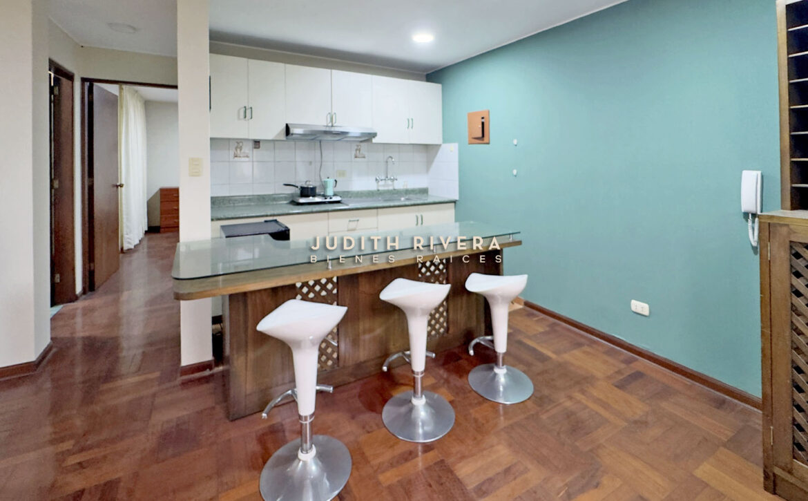 Cocina abierta del departamento en Santiago de Surco, con barra y tres taburetes blancos.
