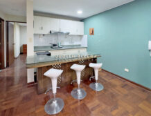 Cocina abierta del departamento en Santiago de Surco, con barra y tres taburetes blancos.