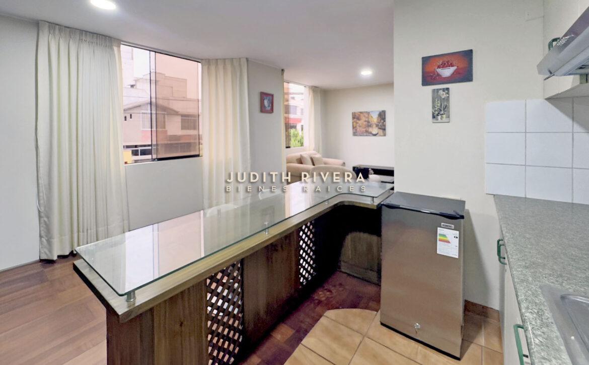 Barra de la cocina abierta hacia la sala, con ventana de cortinas claras y refrigeradora al costado.