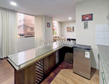 Barra de la cocina abierta hacia la sala, con ventana de cortinas claras y refrigeradora al costado.