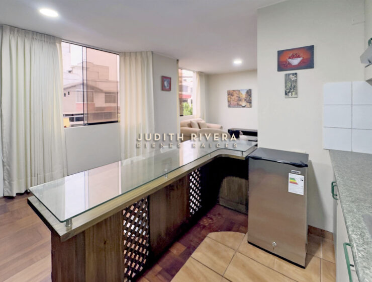 Barra de la cocina abierta hacia la sala, con ventana de cortinas claras y refrigeradora al costado.