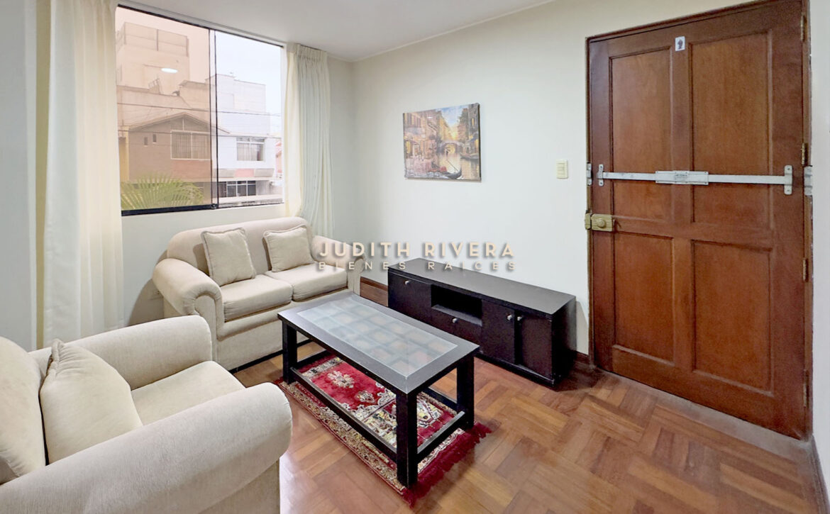 Sala del departamento en Surco con sofás claros y mesa de centro junto a la puerta principal de madera.
