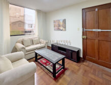 Sala del departamento en Surco con sofás claros y mesa de centro junto a la puerta principal de madera.