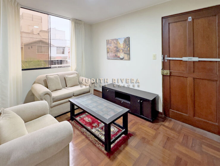Sala del departamento en Surco con sofás claros y mesa de centro junto a la puerta principal de madera.