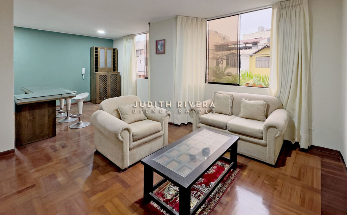 Sala con sofás, mesa de centro y mueble de madera, con ventana a la calle y pared verde agua.