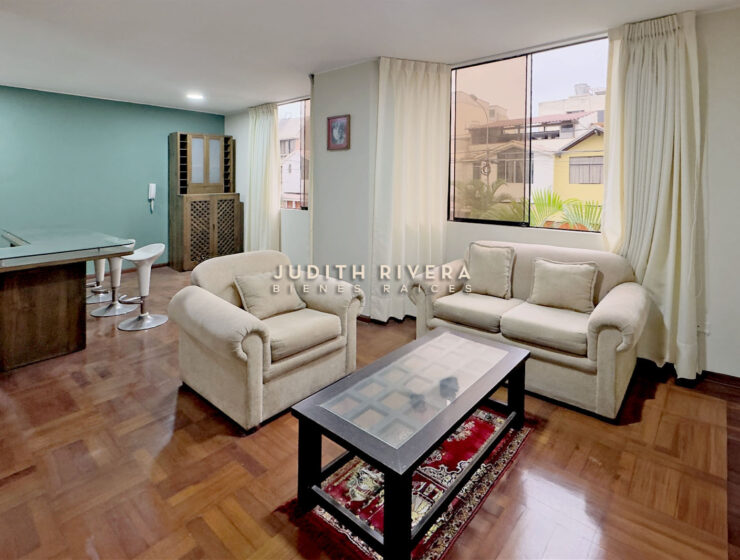 Sala con sofás, mesa de centro y mueble de madera, con ventana a la calle y pared verde agua.