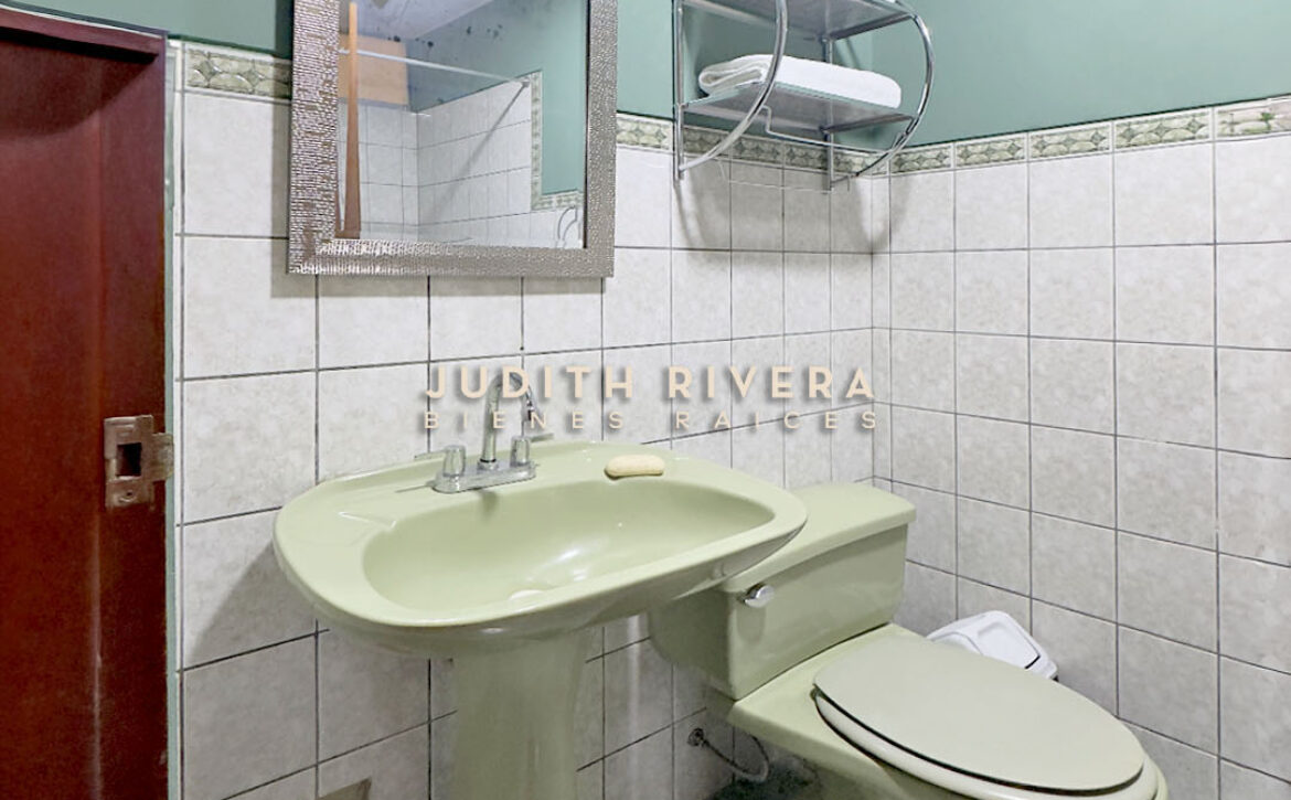Baño completo con lavatorio, espejo y repisas sobre pared de azulejos blancos.