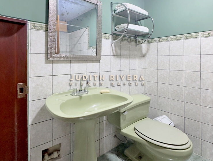 Baño completo con lavatorio, espejo y repisas sobre pared de azulejos blancos.