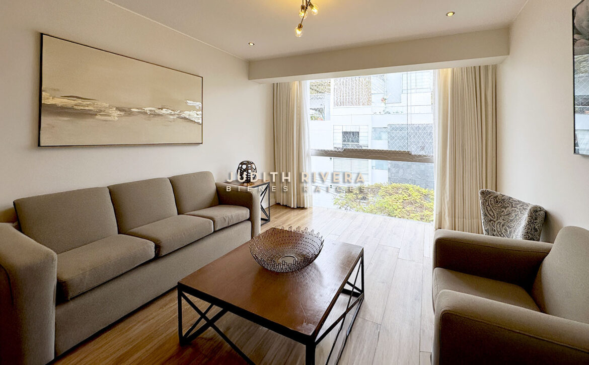 Sala junto al ventanal con sofá largo, mesa de centro y plantas visibles afuera