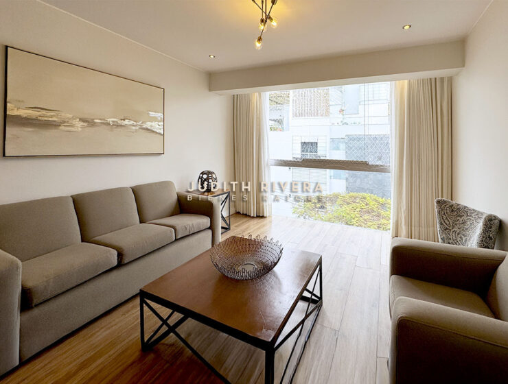 Sala junto al ventanal con sofá largo, mesa de centro y plantas visibles afuera