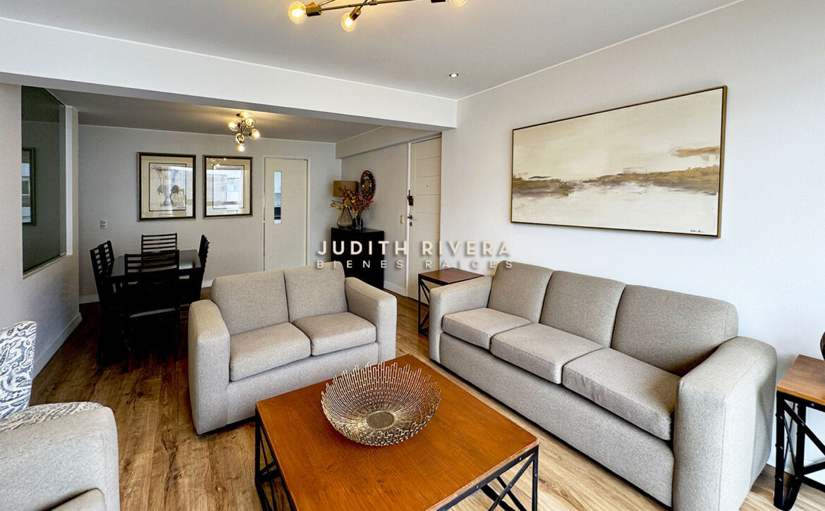 Sala con sofás grises y mesa de centro; al fondo, el comedor y la puerta de ingreso