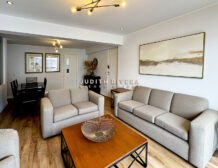 Sala con sofás grises y mesa de centro; al fondo, el comedor y la puerta de ingreso