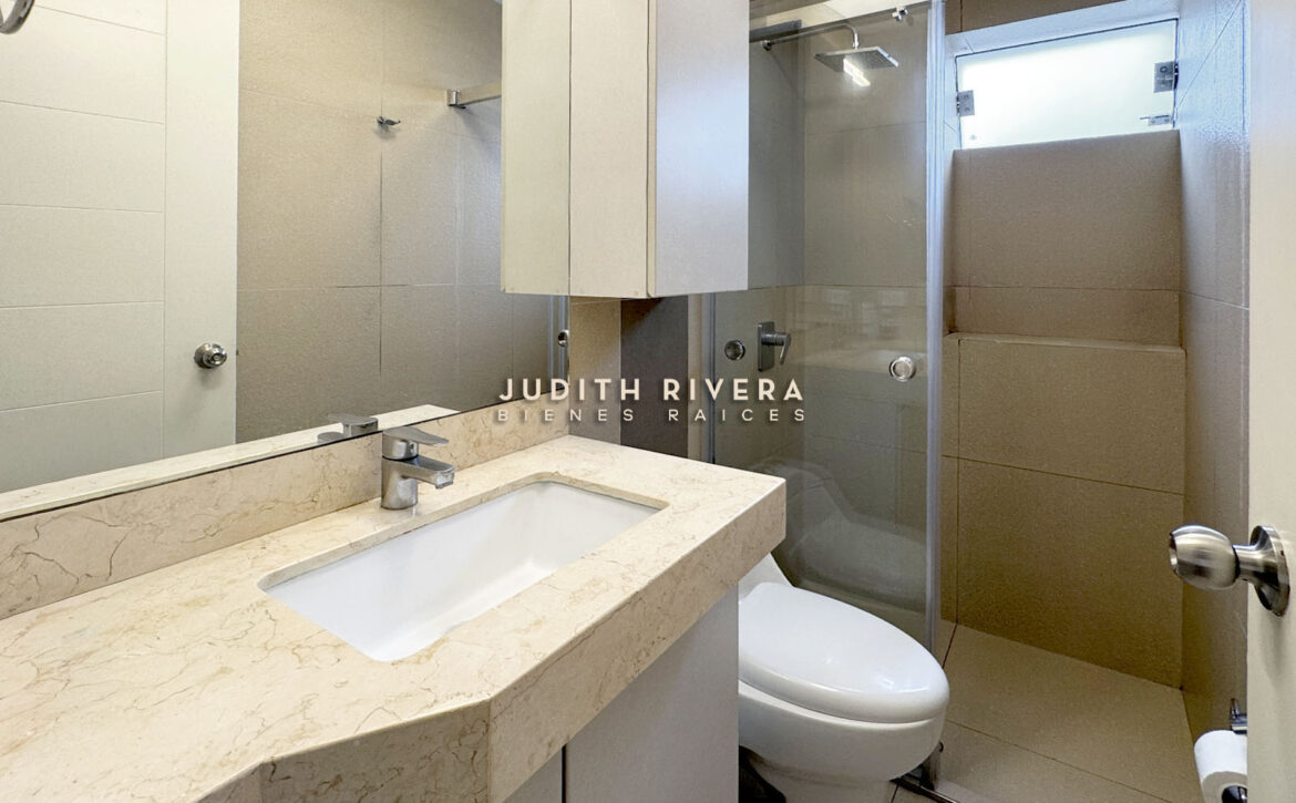 Baño con tablero de mármol claro, ducha con mampara de vidrio y ventana alta